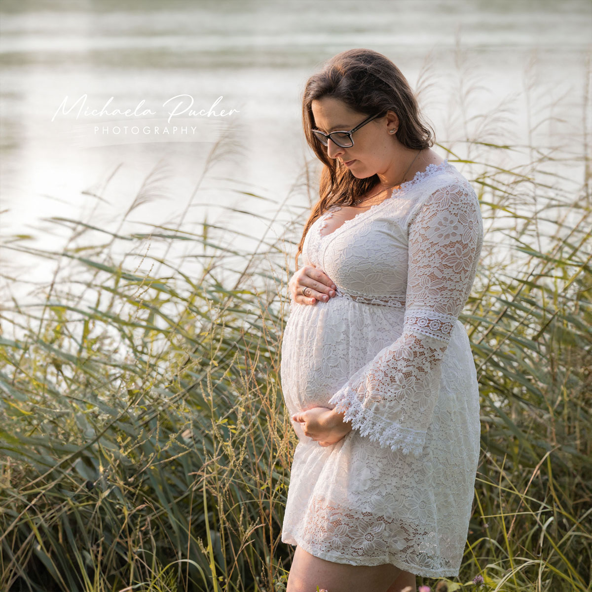 Schwangere Frau in einem weißen Spitzenkleid hält ihren Babybauch, umgeben von hohen Gräsern am Ufer eines Sees bei warmem Abendlicht.